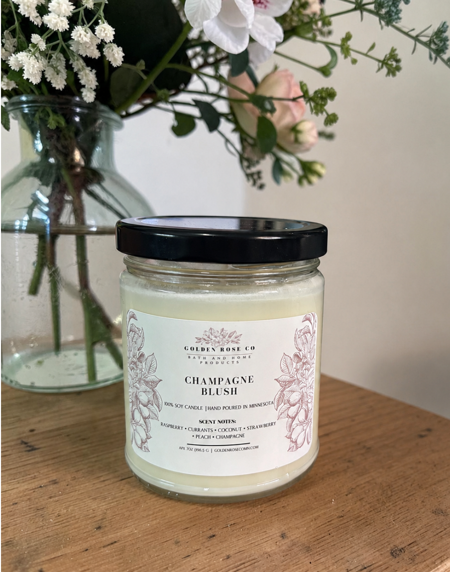 A glass jar candle with a black lid and a cream-colored wax body sits on a wooden table. The label reads 'CHAMPAGNE BLUSH' and shows the brand ' Golden Rose Co