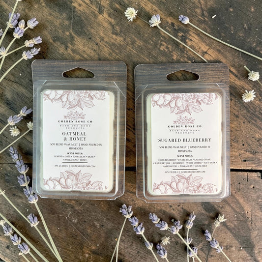 Two packaged soy blend wax melts from Golden Rose Co are arranged side by side on a rustic wooden surface with scattered lavender sprigs.