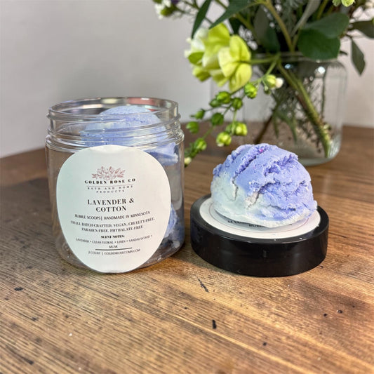 A jar of lavender and cotton bath scoop from Golden Rose Co. sits on a wooden table next to a single scoop of the lavender and cotton bath product on a black lid.