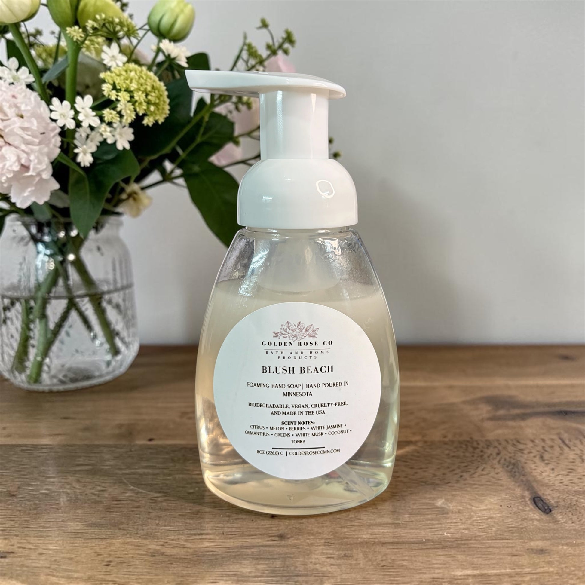A bottle of Golden Rose Co Blush Beach Foaming Hand Soap Poured in Minnesota, featuring a white pump dispenser and a round white label, set on a wooden table with a vase of flowers in the background.